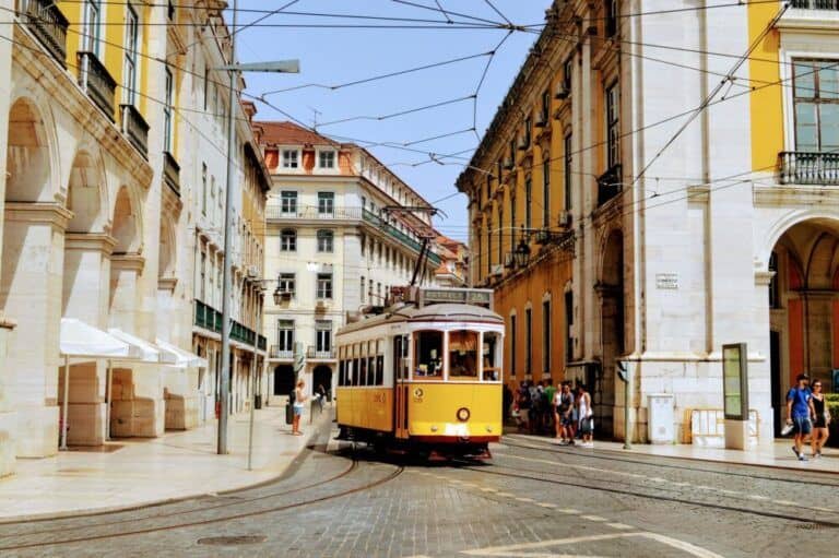 Cartaz Cultural de Lisboa Fotografia de um elétrico amarelo a circular pelas ruas históricas de Lisboa. O elétrico passa entre edifícios antigos com fachadas clássicas, refletindo a arquitetura tradicional da cidade. O céu está limpo e várias pessoas caminham nas proximidades, apreciando o ambiente pitoresco da capital portuguesa.