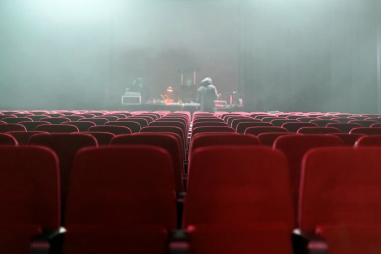 Fotografia de uma sala de teatro vazia com cadeiras vermelhas, enquanto a equipa técnica prepara o palco ao fundo, envolto em névoa. A imagem é ideal para promover teatro infantil em Lisboa, destacando a preparação e a antecipação antes do início de uma peça, criando um ambiente mágico e envolvente.