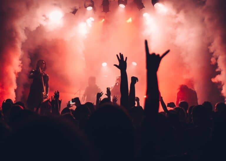 Fotografia de um concerto ao vivo em Lisboa, com uma multidão animada a levantar as mãos em frente ao palco iluminado por luzes vermelhas e névoa. A imagem capta a energia e a emoção do evento, sendo ideal para promover concertos em Lisboa, destacando a vibrante cena musical da cidade.