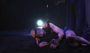 Fotografia de uma performance teatral intensa em Lisboa, mostrando um ator coberto de terra e ligado a um arnês, deitado no chão sob uma luz dramática. A imagem captura a emoção e a dedicação dos artistas, sendo ideal para promover peças de teatro em Lisboa, destacando a diversidade e a profundidade das produções teatrais na cidade.