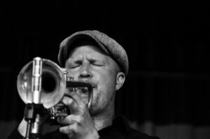 Fotografia a preto e branco de um músico de jazz a tocar trompete com paixão. O músico, usando um boné, é captado a meio de uma atuação com os olhos fechados, transmitindo profunda emoção e concentração. Os detalhes do trompete são proeminentes em primeiro plano, com um suporte de microfone visível, sugerindo um cenário de performance ao vivo Jazz em Lisboa: Os melhores sítios para ouvir jazz ao vivo