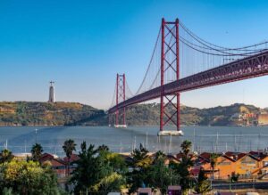 Cartaz Cultural de Lisboa "Fotografia da Ponte 25 de Abril em Lisboa, com o Cristo Rei visível ao fundo. A imagem mostra um céu azul claro, o rio Tejo e a marina em primeiro plano, cercada por vegetação e edifícios. Ideal para destacar atividades e atrações turísticas em Lisboa, capturando a beleza icónica da cidade.