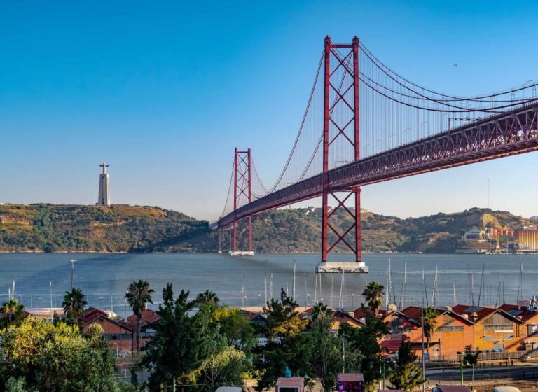 Cartaz Cultural de Lisboa "Fotografia da Ponte 25 de Abril em Lisboa, com o Cristo Rei visível ao fundo. A imagem mostra um céu azul claro, o rio Tejo e a marina em primeiro plano, cercada por vegetação e edifícios. Ideal para destacar atividades e atrações turísticas em Lisboa, capturando a beleza icónica da cidade.