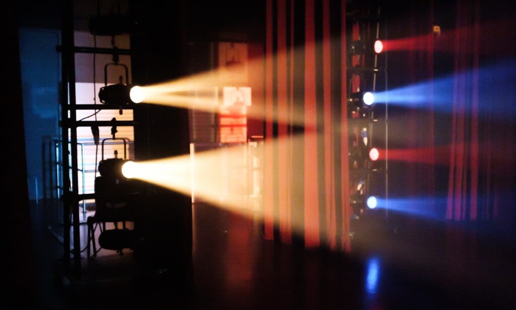 Fotografia de luzes de palco coloridas a iluminar uma cena teatral em Lisboa. A imagem destaca os feixes de luz nas cores azul, vermelho e amarelo, criando uma atmosfera dramática. Ideal para promover peças de teatro em Lisboa, destacando a criatividade e a produção técnica das apresentações teatrais na cidade.
