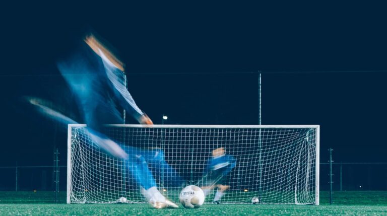 "Fotografia de um jogador de futebol a chutar uma bola em direção à baliza, capturada em movimento, com a rede da baliza ao fundo durante a noite. A imagem é ideal para ilustrar o tema 'Onde dá a Bola - Hoje', destacando o calendário de jogos de futebol na TV portuguesa e ajudando os fãs a acompanhar as partidas ao vivo.