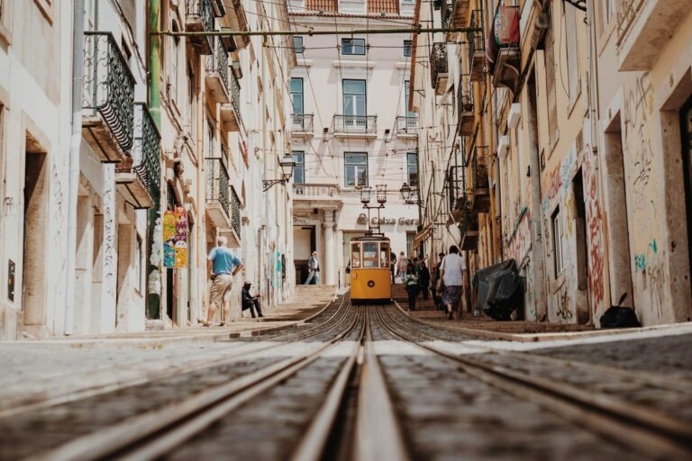 Fotografia de um elétrico amarelo a subir uma rua inclinada em Lisboa, rodeado por edifícios históricos e pessoas a passear. A imagem capta a essência da cidade com suas ruas pitorescas e atmosfera vibrante, sendo ideal para destacar atividades e passeios a realizar este fim de semana em Lisboa.