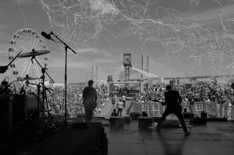 Fotografia em preto e branco de um concerto ao ar livre em Lisboa, com uma banda a atuar no palco e uma grande multidão a assistir. Ao fundo, é visível uma roda-gigante e uma ponte, criando uma atmosfera vibrante de festival. Ideal para promover concertos em Lisboa, capturando a energia e a emoção de eventos musicais ao vivo na cidade.