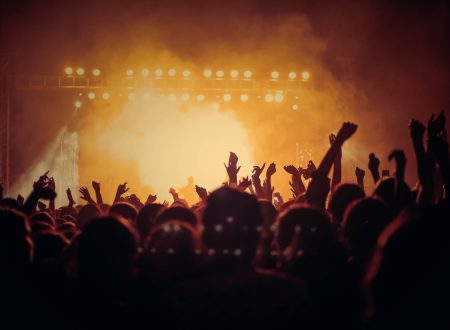 Cartaz Cultural de Lisboa Fotografia de um concerto ao vivo com uma multidão de pessoas a levantar as mãos em frente a um palco iluminado com luzes quentes. O ambiente é vibrante e cheio de energia, capturando a essência de uma experiência musical ao vivo. As luzes e a fumaça no palco criam uma atmosfera intensa e envolvente