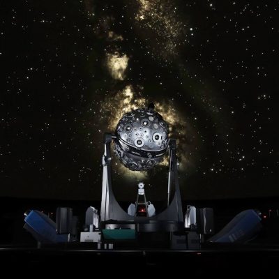 Fotografia do interior do Planetário Calouste Gulbenkian em Lisboa, mostrando um projetor de estrelas em frente a uma projeção do céu noturno repleto de estrelas. A imagem captura a atmosfera imersiva e educativa do planetário, ideal para promover visitas e eventos no Planetário Calouste Gulbenkian em Lisboa.
