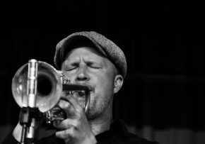 Fotografia a preto e branco de um músico de jazz a tocar trompete com paixão. O músico, usando um boné, é captado a meio de uma atuação com os olhos fechados, transmitindo profunda emoção e concentração. Os detalhes do trompete são proeminentes em primeiro plano, com um suporte de microfone visível, sugerindo um cenário de performance ao vivo Jazz em Lisboa: Os melhores sítios para ouvir jazz ao vivo