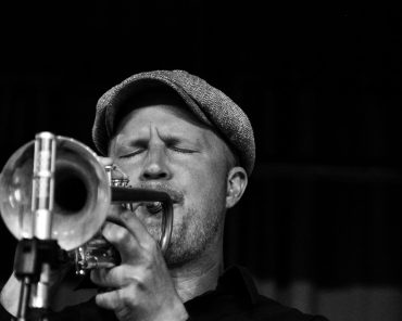 Fotografia a preto e branco de um músico de jazz a tocar trompete com paixão. O músico, usando um boné, é captado a meio de uma atuação com os olhos fechados, transmitindo profunda emoção e concentração. Os detalhes do trompete são proeminentes em primeiro plano, com um suporte de microfone visível, sugerindo um cenário de performance ao vivo Jazz em Lisboa: Os melhores sítios para ouvir jazz ao vivo