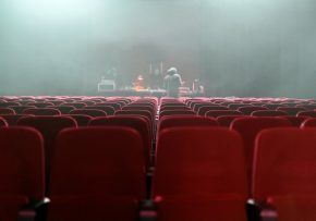 Fotografia de uma sala de teatro vazia com cadeiras vermelhas, enquanto a equipa técnica prepara o palco ao fundo, envolto em névoa. A imagem é ideal para promover teatro infantil em Lisboa, destacando a preparação e a antecipação antes do início de uma peça, criando um ambiente mágico e envolvente.