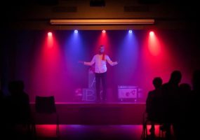 Fotografia de um comediante a realizar um espetáculo de stand-up comedy em Lisboa, iluminado por luzes coloridas vermelhas e azuis. A imagem capta a atmosfera vibrante e envolvente do evento, ideal para promover noites de stand-up comedy em Lisboa, destacando a diversão e o entretenimento disponíveis na cidade.