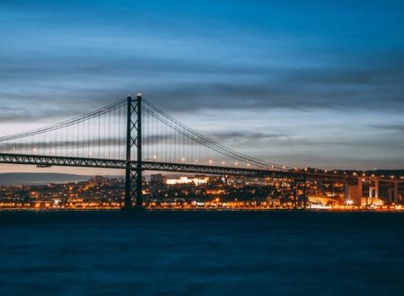 Fotografia noturna da Ponte 25 de Abril em Lisboa, iluminada pelas luzes da cidade refletidas no rio Tejo. A imagem destaca a beleza da cidade à noite, sendo ideal para promover atividades noturnas em Lisboa, como passeios, eventos e a vibrante vida noturna